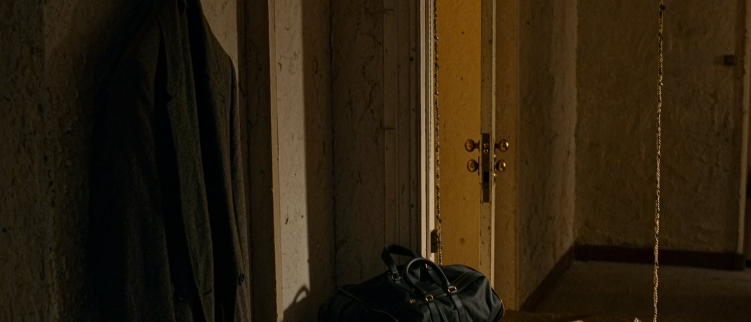 a hall seen close with a coat on the hook, a packed bag by the door and a folded decree printed from the gazette on the console, the door with its chain soft behind