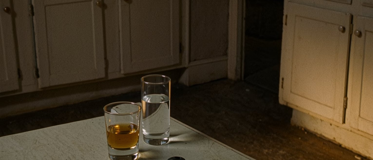 a kitchen table seen close with two glasses, one of whisky and one of water, and a single house key set between them, the kettle, a bowl of fruit and the worktop close behind