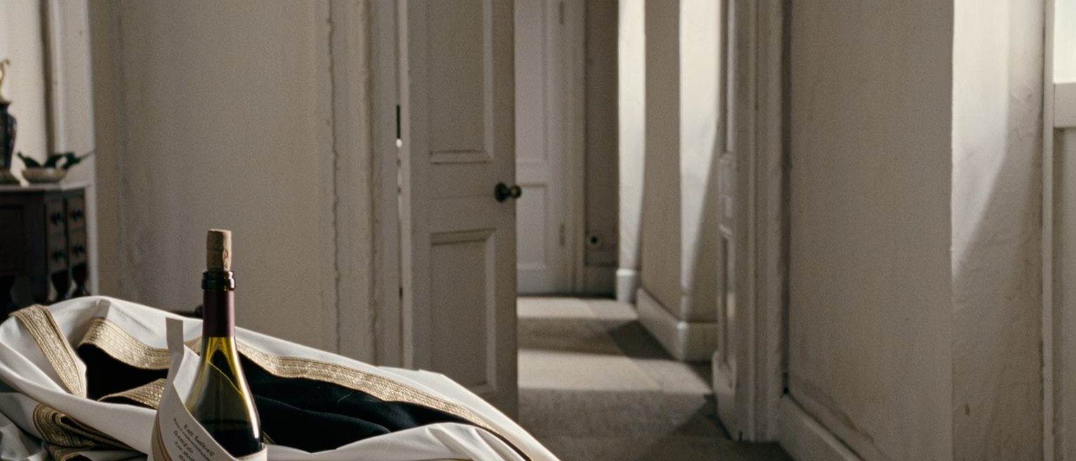 a flat's hall seen close with an envelope from the registry in a dish beside a set of keys and a bottle of wine still in its paper bag, a folded abaya on the console, the door soft behind