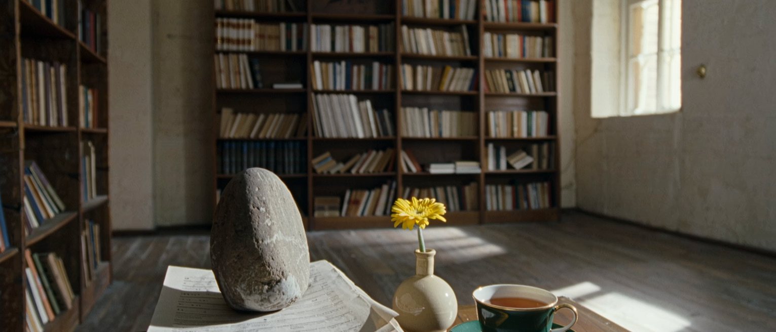 a study seen close with a stack of typed pages weighted by a stone, a vase with one yellow flower, a pencil sharpened and a cup of tea, the bookshelves close behind