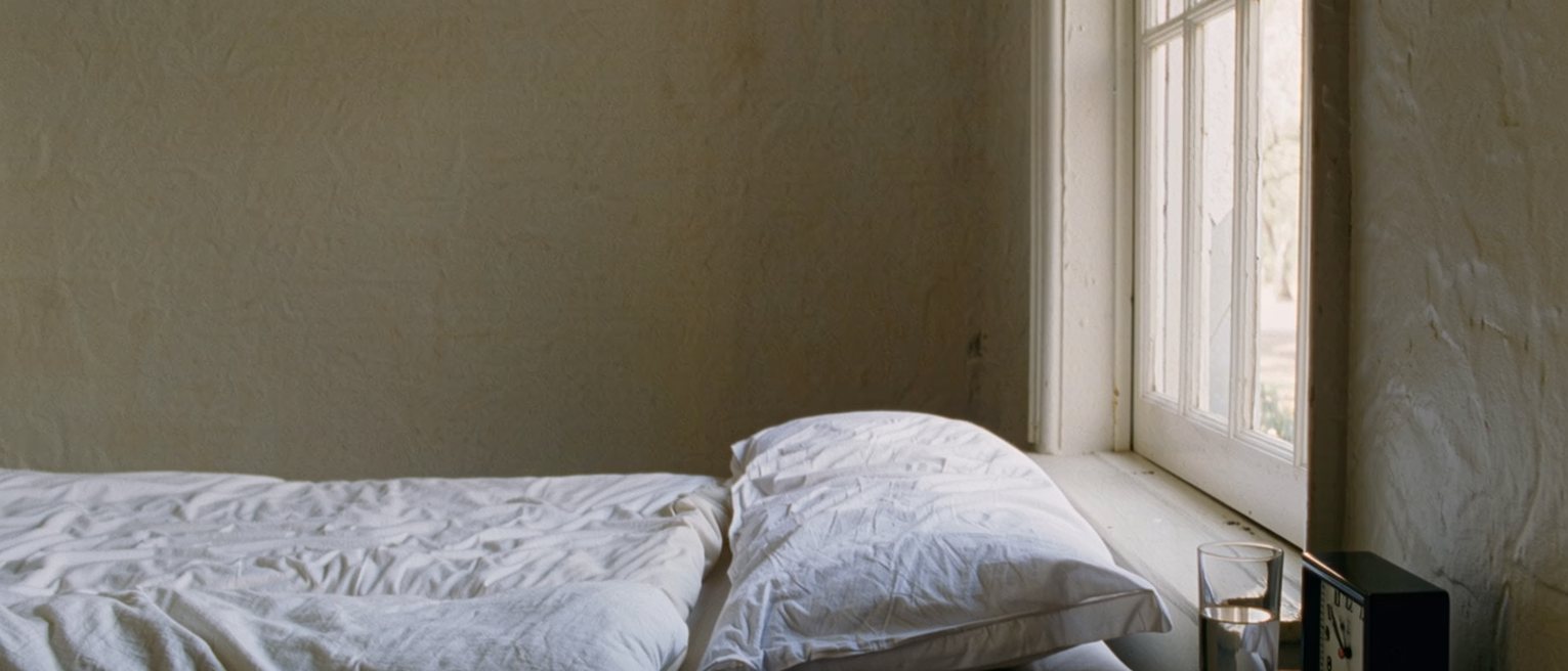 a bedroom seen close with a folded sheet of paper with a numbered list written on it laid on the bedside table beside a room key on its fob, a glass of water and a small clock, the bed with its cover turned back close behind and the tall window soft beyond
