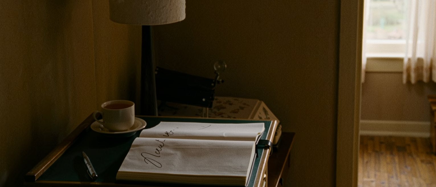 a writing desk seen close with a notebook open at a page where one verb is written and underlined, a pen laid down, a cup of tea, the lamp close behind and the window soft beyond
