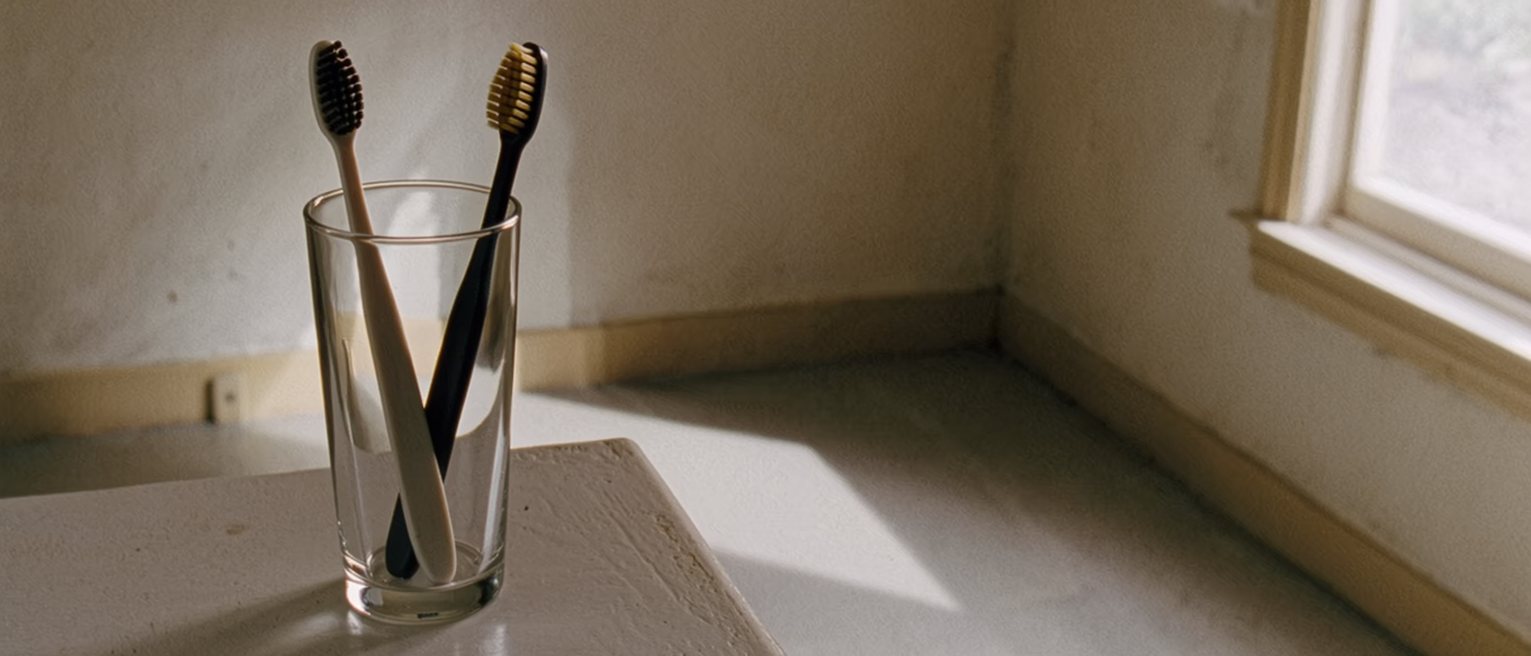 a bathroom shelf with two toothbrushes standing in one glass and nothing else on it