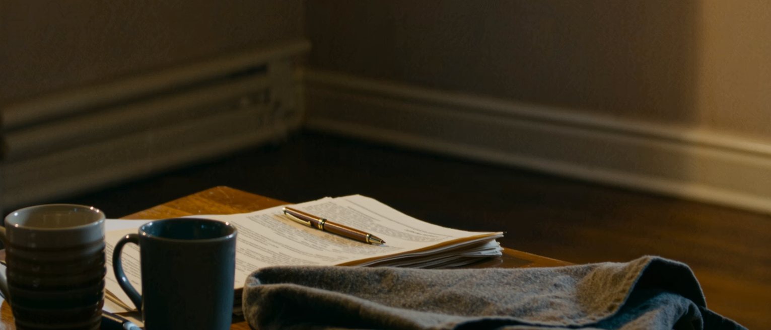 a kitchen table with a folded document laid square between two cups and a pen set across it, an apron folded beside them