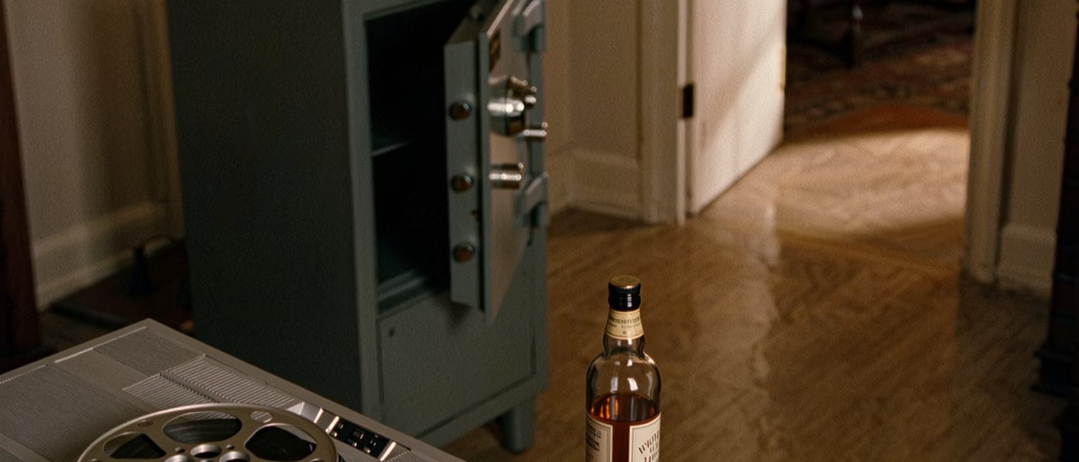 a study seen close with a film reel in its tin on the desk, a projector unplugged, a bottle of whisky and two glasses, a safe with its door ajar soft behind
