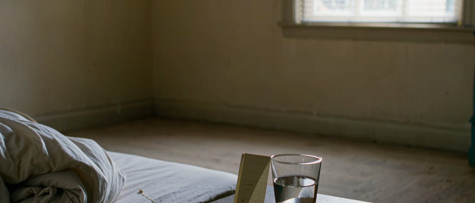 a bedside table with a folded note propped against a glass of water, the covers turned down on one side