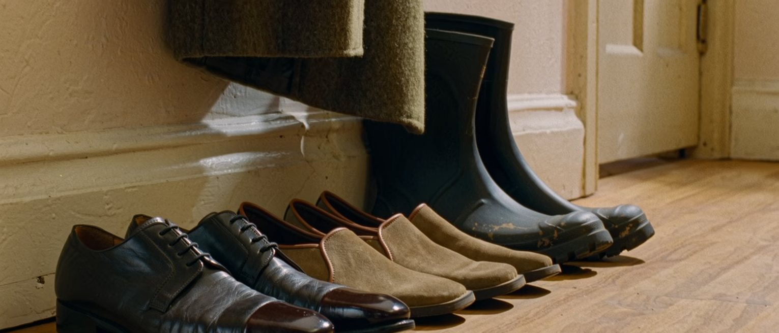 a hall seen close with three pairs of shoes in a row, each for a different life, and a coat half taken off the hook, the door on the latch behind