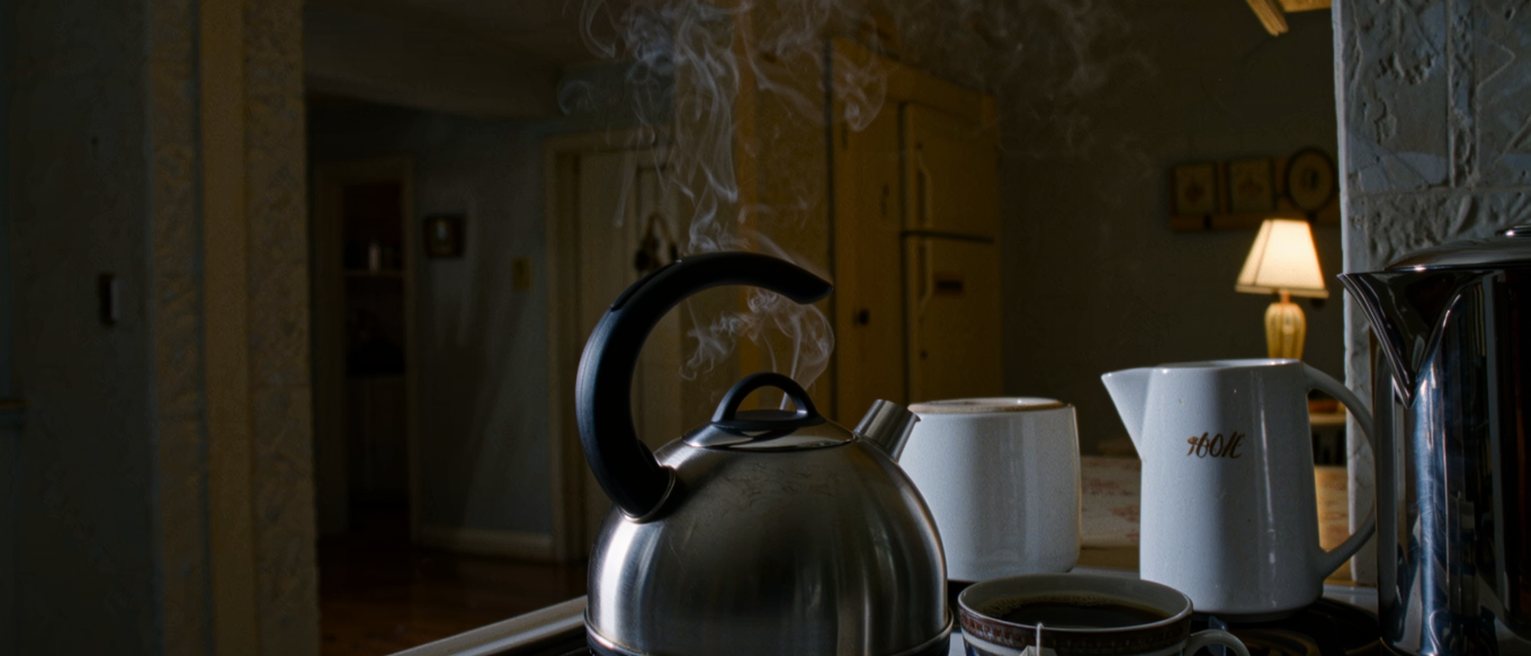 a kitchen worktop seen close with the kettle boiled and left, a cup with the teabag still in it and a pot of coffee on the stove, both untouched, the toaster, a bowl of fruit and the shelves of jars close behind
