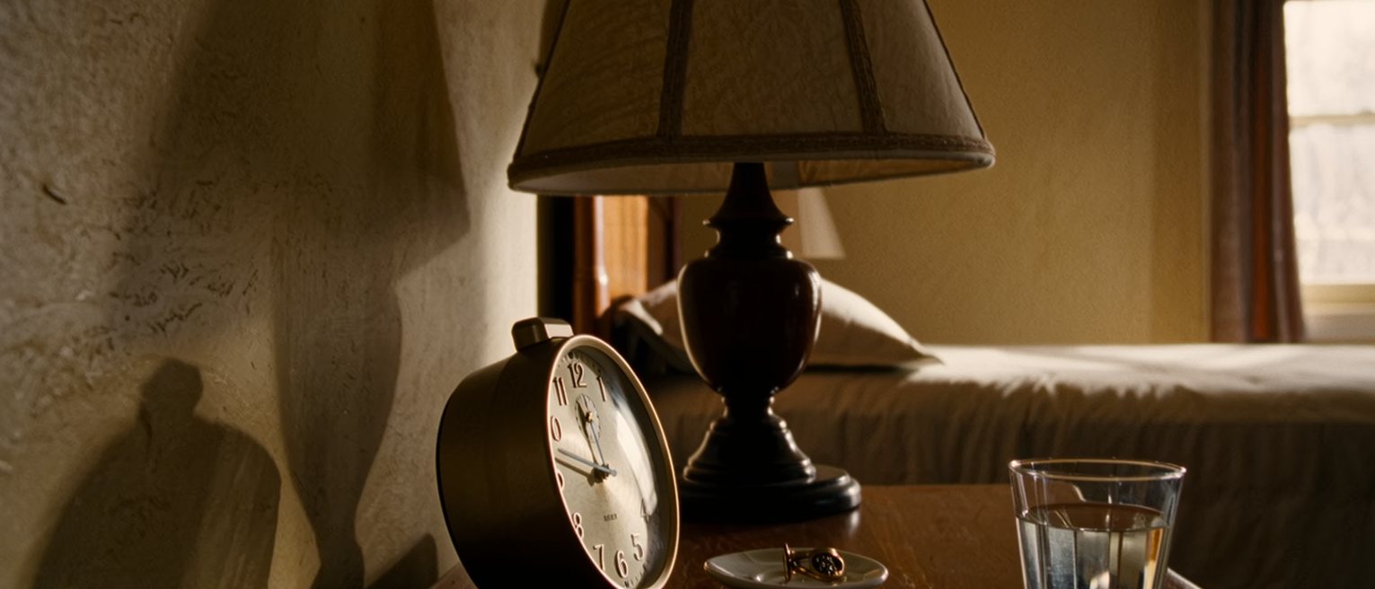 a bedside table seen close with a travel clock turned to face the wall, a single earring in a saucer and a glass of water half drunk, the lamp and the turned-down bed close behind