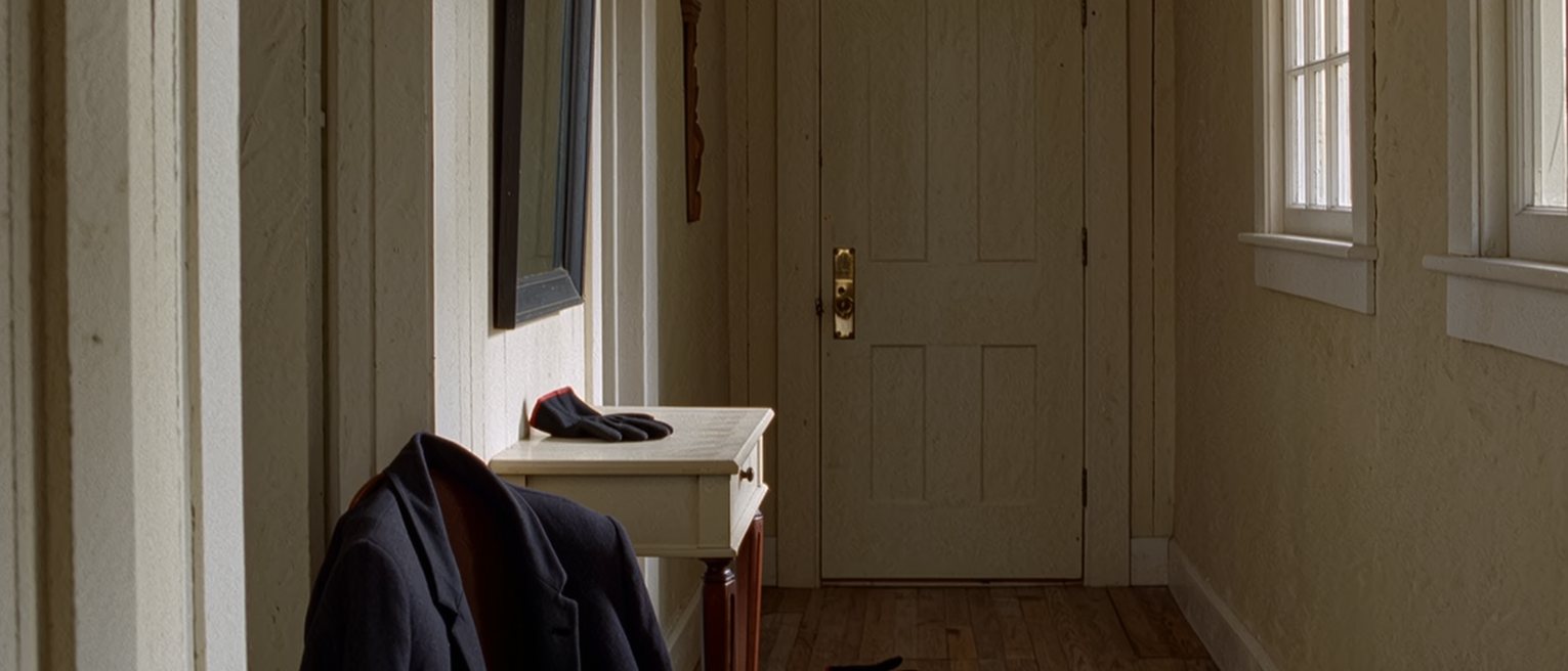 a hall seen close with a coat laid ready over the chair, one glove on the console and the other on the floor beneath it, the front door and its brass latch close behind