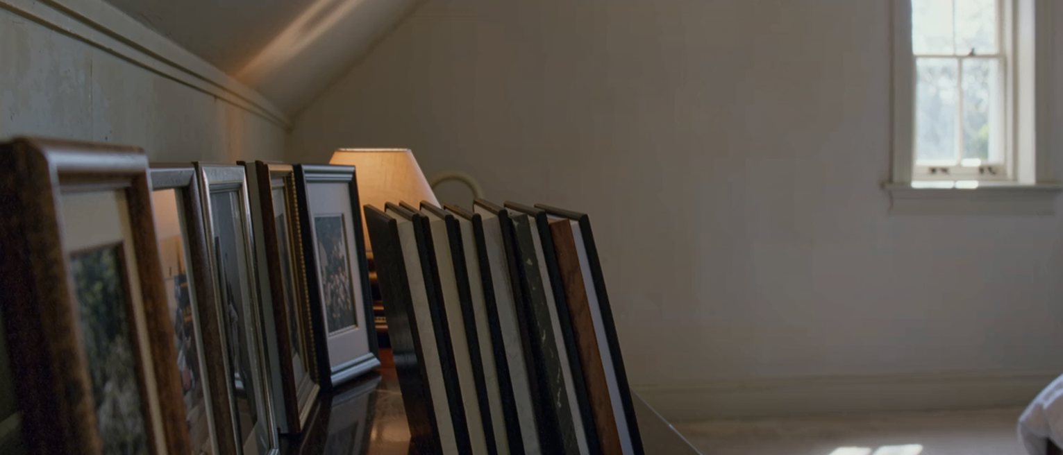 a bedroom dresser with a row of framed photographs turned face down, all the frames the same size, a set of keys and a lipstick beside them and the lamp soft behind