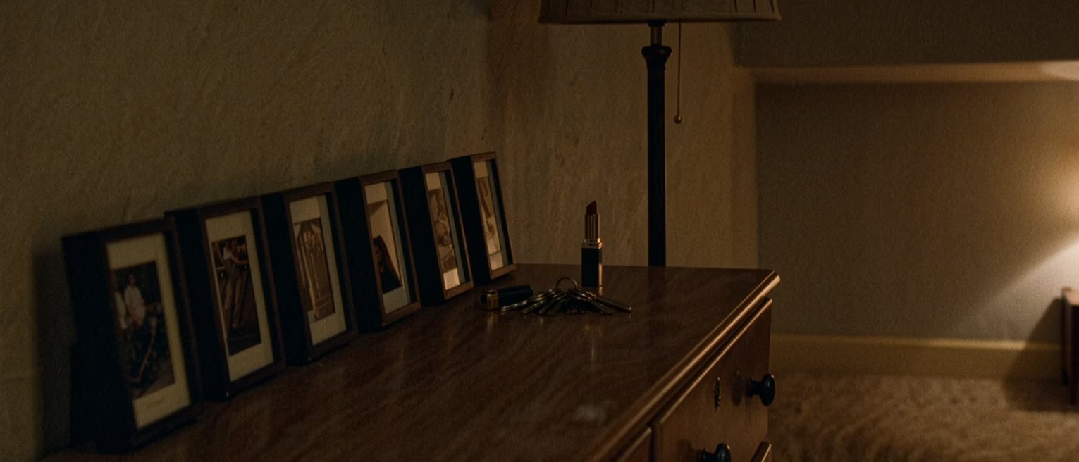 a bedroom dresser with a row of framed photographs turned face down, all the frames the same size, a set of keys and a lipstick beside them and the lamp soft behind